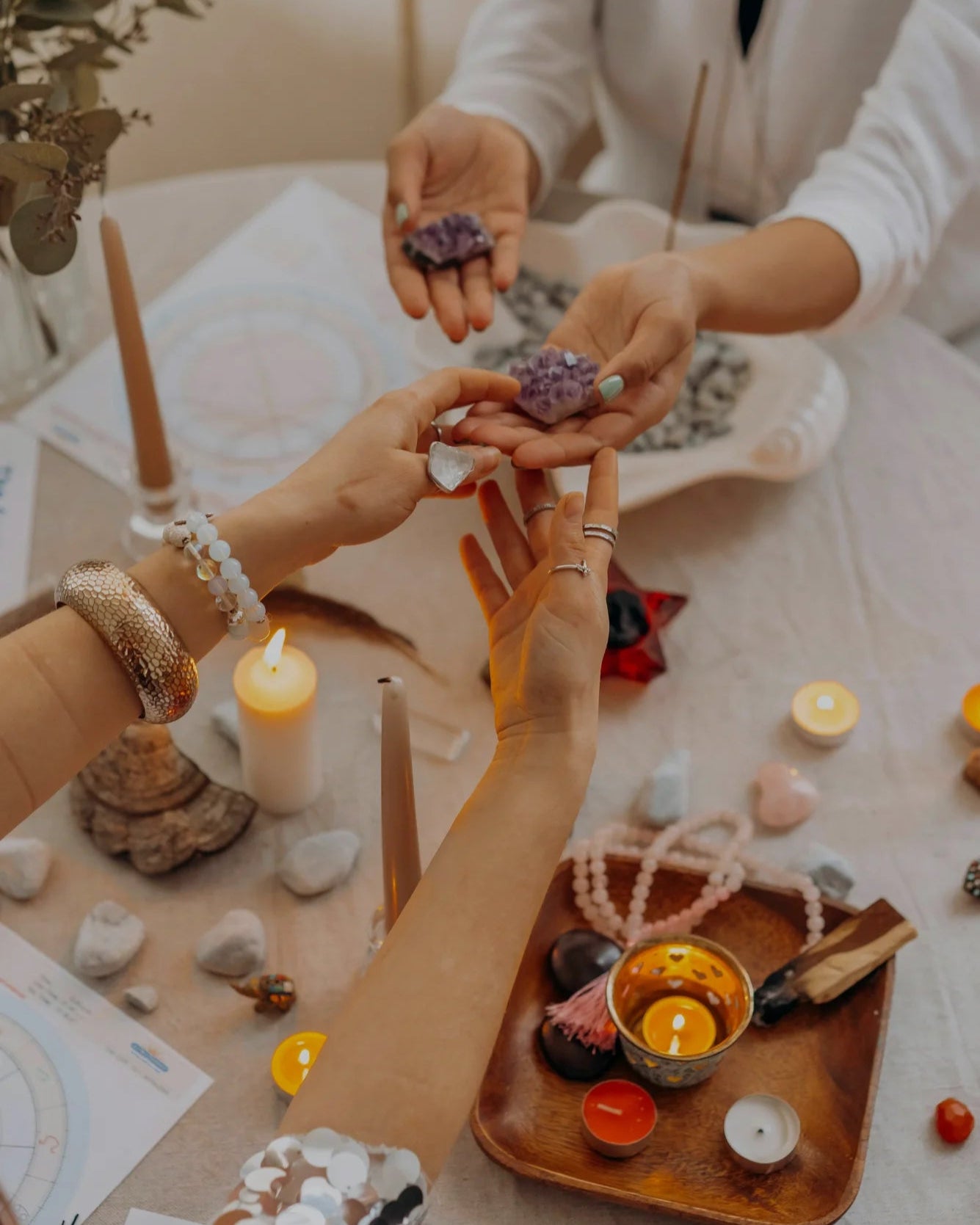 Two people holding small purple stones over a table with candles, crystals, and other objects.